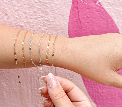 Close-up of a wrist stacked with delicate gold, rose gold and silver chain bracelets against a pink textured wall, another hand with floral nail art adjusting a chain.