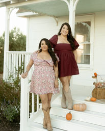 Two women in floral and burgundy dresses with cowboy boots posing on a white farmhouse-style porch decorated with pumpkins and a wicker basket for a fall fashion photo