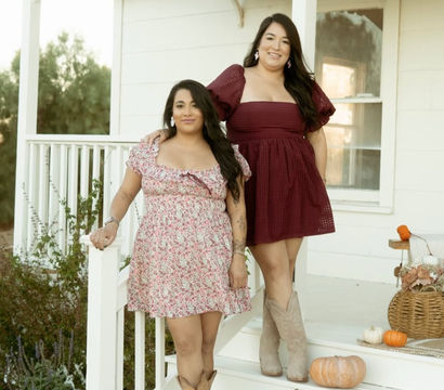 Two women in floral and burgundy dresses with cowboy boots posing on a white farmhouse-style porch decorated with pumpkins and a wicker basket for a fall fashion photo