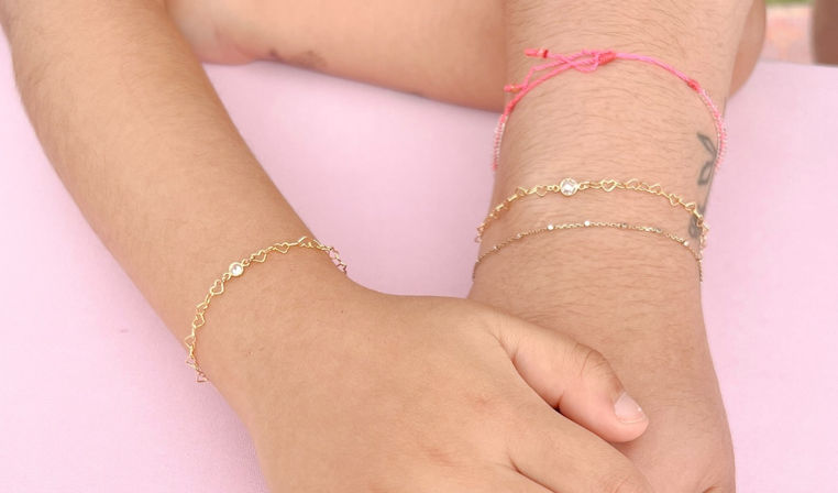 Close-up of two wrists with delicate gold heart-link and thin chain bracelets plus a pink string bracelet, stacked on a soft pink background, jewel-accented and minimalist style