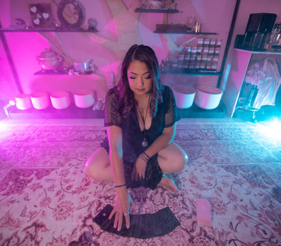 Person kneeling on a patterned rug in a pink-and-blue neon-lit boho studio, arranging a fanned tarot card spread with shelves of jars and bowls in the background.