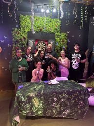 Eight people posing in a festive indoor game-room setting in front of a leafy green wall and dartboard, holding small jars and smiling around a tropical-print table under hanging tinsel and butterfly decorations.