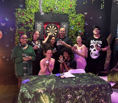 Eight people posing in a festive indoor game-room setting in front of a leafy green wall and dartboard, holding small jars and smiling around a tropical-print table under hanging tinsel and butterfly decorations.