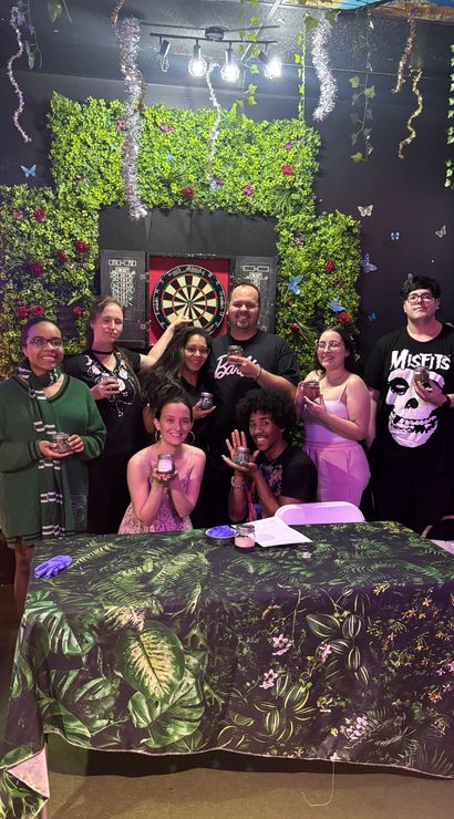 Eight people posing in a festive indoor game-room setting in front of a leafy green wall and dartboard, holding small jars and smiling around a tropical-print table under hanging tinsel and butterfly decorations.