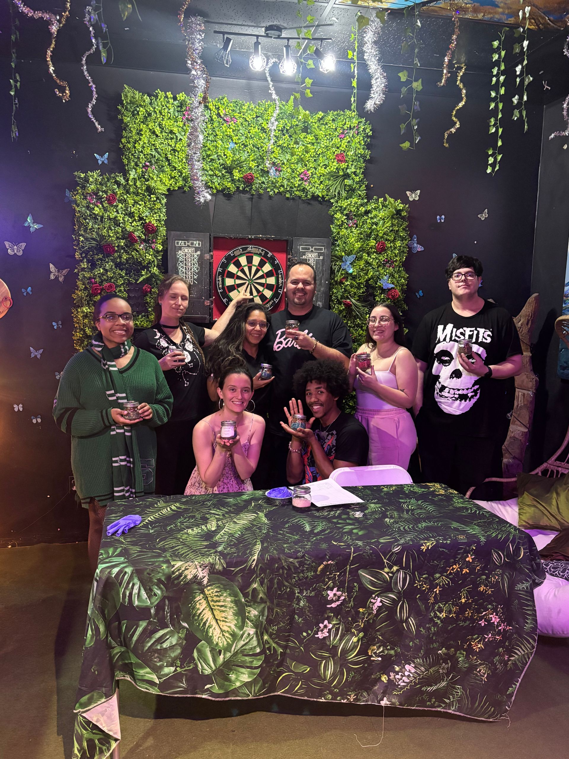 Eight people posing in a festive indoor game-room setting in front of a leafy green wall and dartboard, holding small jars and smiling around a tropical-print table under hanging tinsel and butterfly decorations.