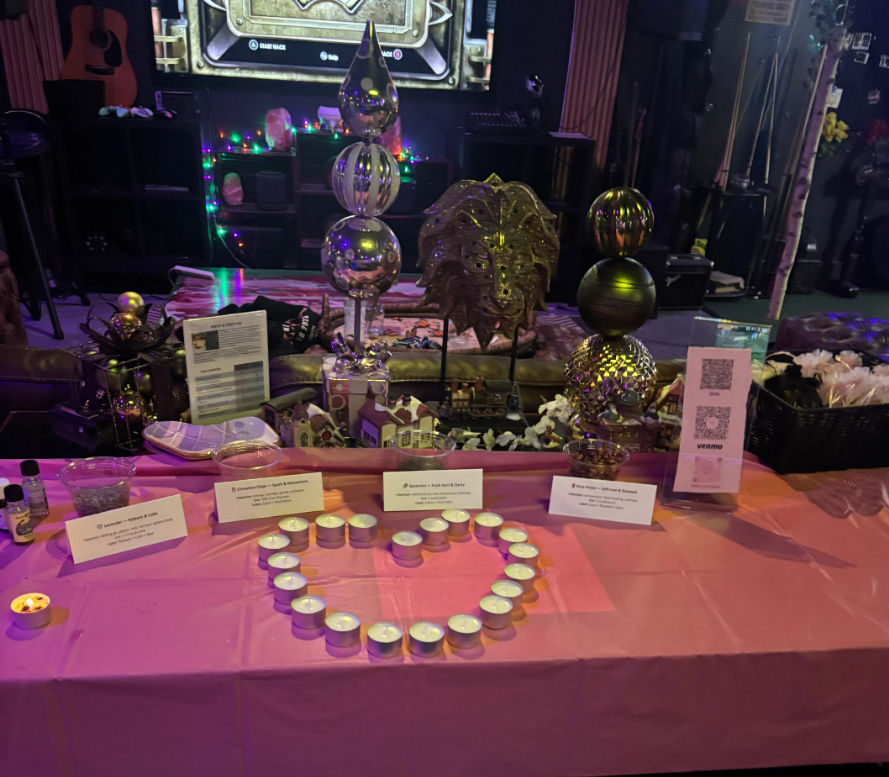 Festive indoor event table display on a pink tablecloth with tea light candles arranged in a heart, decorative metal orb sculptures, miniature model houses, informational cards and a QR code, purple string lights and stage backdrop.