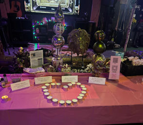 Festive indoor event table display on a pink tablecloth with tea light candles arranged in a heart, decorative metal orb sculptures, miniature model houses, informational cards and a QR code, purple string lights and stage backdrop.