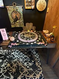 Cozy spiritual reading table with a black astrology tarot cloth showing a zodiac wheel, crystal ball, candles and trinkets, raven tapestry and rune drum on a moody boho wall.