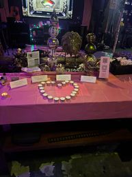 Sparkly indoor event table with tealight candles arranged in a heart on a pink tablecloth, surrounded by metallic orb decorations, small signs, a QR code and fairy lights