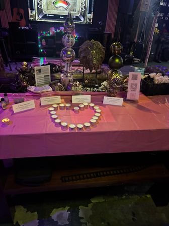 Sparkly indoor event table with tealight candles arranged in a heart on a pink tablecloth, surrounded by metallic orb decorations, small signs, a QR code and fairy lights