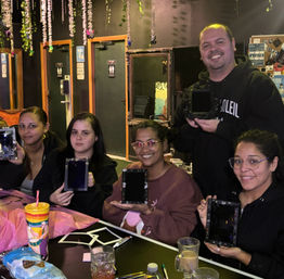 Five people smiling at an indoor craft workshop, seated around a decorated table holding small DIY framed mirrors, with hanging floral vines, drinks and art supplies visible.