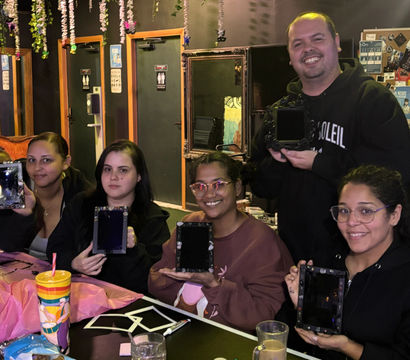 Five people smiling at an indoor craft workshop, seated around a decorated table holding small DIY framed mirrors, with hanging floral vines, drinks and art supplies visible.
