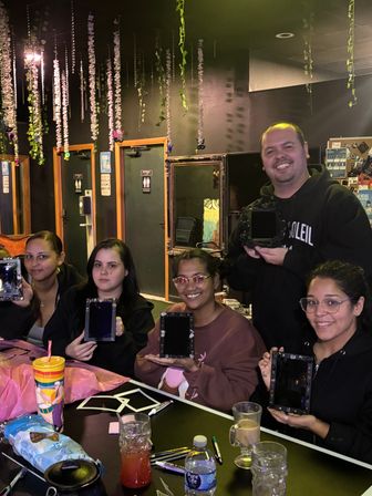 Five adults at a cozy indoor DIY craft night in a craft studio, smiling and holding decorated black shadowbox frames beneath hanging floral garlands, with drinks and snacks on the table.