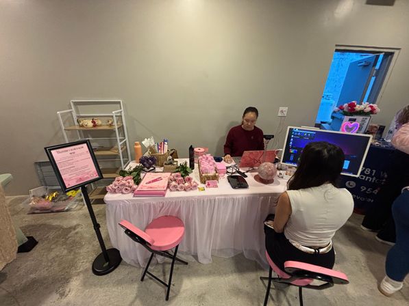 Pink-themed pop-up booth at an indoor market: vendor behind a table with laptop, monitor and crystal ball, stacks of pink roses, brochures and two pink chairs.