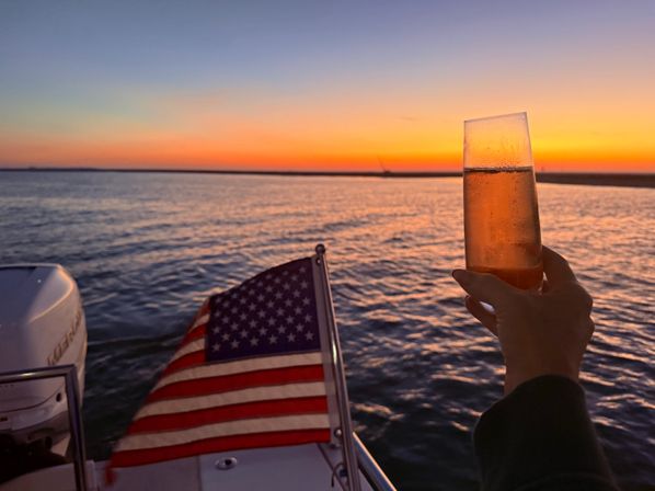 Sunset cruise on calm water—hand holding a chilled champagne flute aboard a boat stern with a waving American flag against an orange-pink horizon.