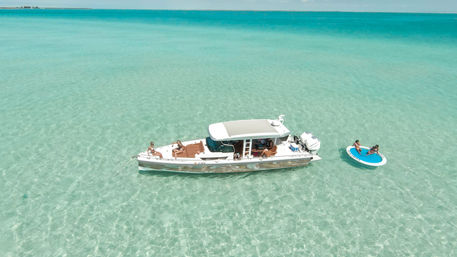 Aerial view of a luxury motorboat anchored in crystal-clear shallow turquoise tropical water, sun-soaked passengers lounging on deck and two people relaxing on a round inflatable swim platform
