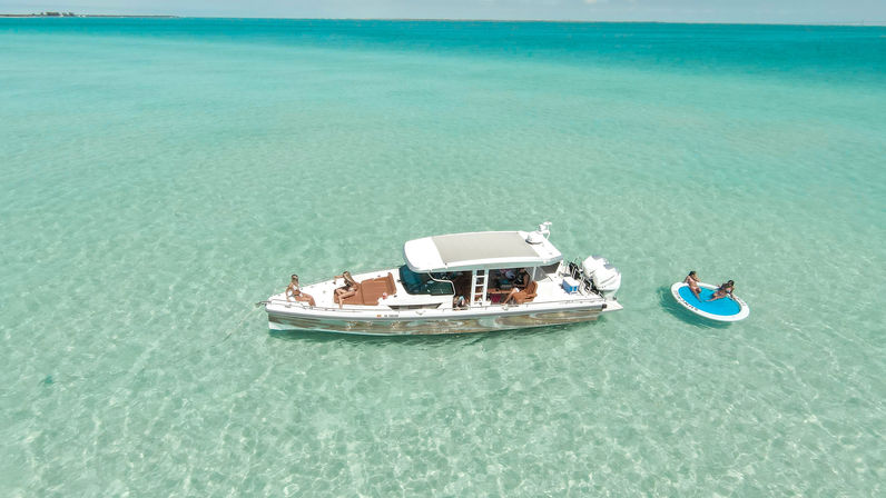 Aerial view of a luxury motorboat anchored in crystal-clear shallow turquoise tropical water, sun-soaked passengers lounging on deck and two people relaxing on a round inflatable swim platform