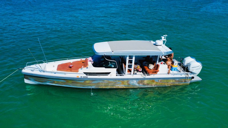 Aerial shot of a sleek center-console motorboat with twin outboard engines anchored in clear turquoise water, sun-lounges and a person on the stern enjoying a sunny day.