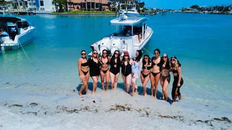 Group of women in swimsuits posing on a sandy shore by a white powerboat in clear turquoise coastal water, sunny waterfront homes and docks in the background.