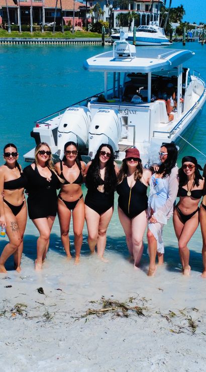 Group of women in swimsuits posing on a sandy shore by a white powerboat in clear turquoise coastal water, sunny waterfront homes and docks in the background.