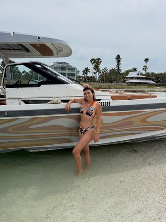 Smiling person in a colorful bikini standing in shallow turquoise water leaning against a moored motorboat with a wavy tan-and-gray hull, palm trees and coastal homes on a tropical shoreline under a cloudy sky.