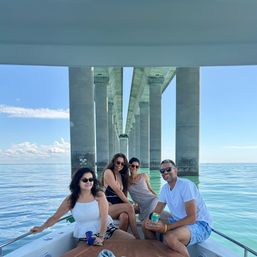 Four friends relaxing on a small boat beneath a long concrete bridge with towering pillars, calm turquoise water, clear blue sky, sunglasses and drinks on a sunny summer outing