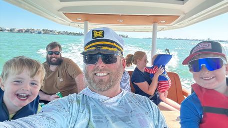 Smiling family on a sunny boat ride over turquoise coastal waters — man in a captain's hat taking a selfie with kids in life jackets and a woman holding a baby, shoreline homes in the distance.