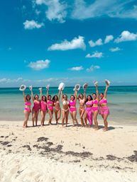 Twelve women in pink swimsuits (one in white) standing on a tropical sandy beach with turquoise water and clear blue sky, smiling and raising patterned sun hats in celebration.