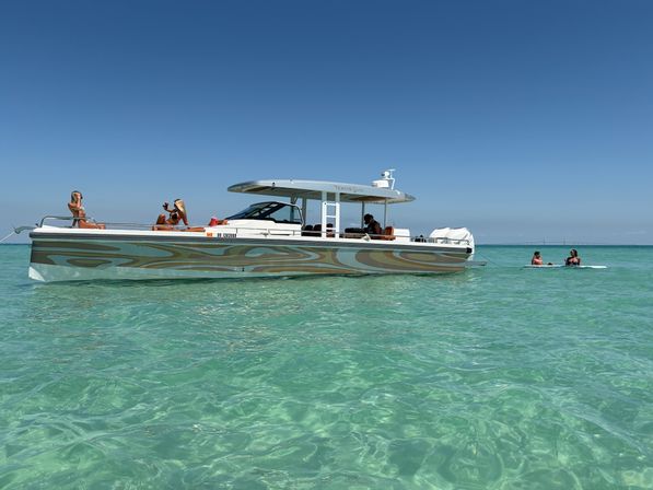 Sunlit motorboat anchored in shallow crystal-clear turquoise coastal waters with two people sunbathing on deck and two paddleboarding nearby under a cloudless blue sky