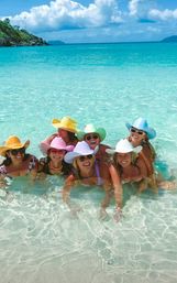 Seven friends in colorful cowboy hats and sunglasses laughing in shallow, crystal-clear turquoise water on a sunny tropical beach with white sand, blue sky and distant island.