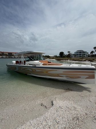 Sleek tan-and-white powerboat with cushioned bow and T-top beached in shallow clear turquoise water along a sandy shell beach, palm trees and modern waterfront homes visible under an overcast sky