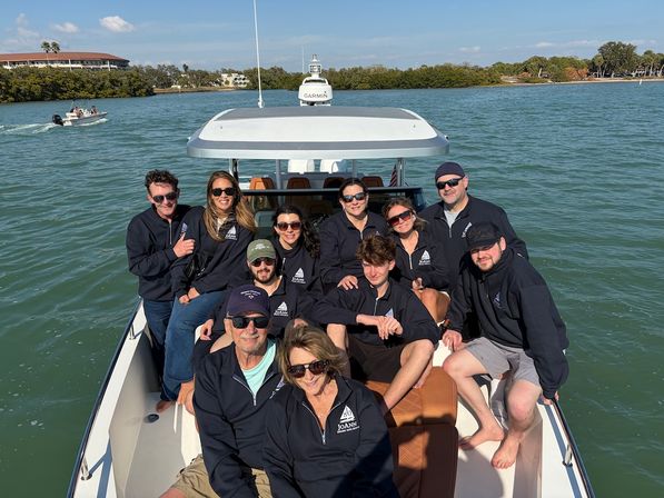 Group of people wearing matching navy jackets smiling and posing on a motorboat in calm blue-green coastal waters on a sunny day with shoreline and mangroves in the background.