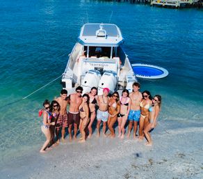 A dozen friends in swimsuits posing on a sandy beach beside an anchored white powerboat in clear turquoise shallow water with a circular inflatable water trampoline nearby.