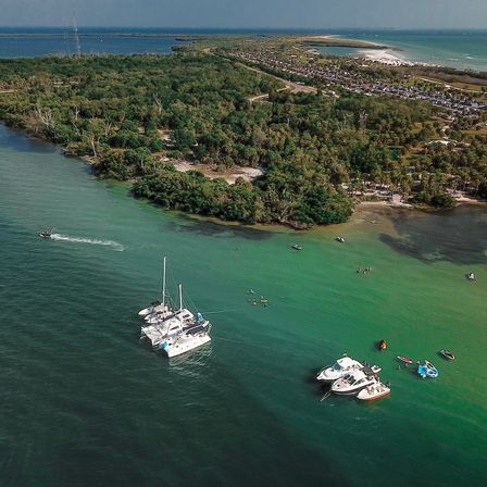 Aerial view of a sunlit barrier island and turquoise bay with anchored catamaran and yachts, kayakers and small boats near a sandy beach and tree-lined shoreline