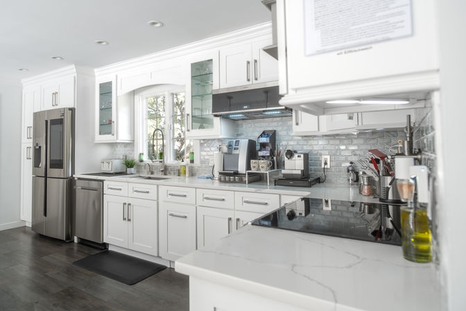 Bright modern white kitchen with shaker cabinets, stainless-steel French-door refrigerator and dishwasher, quartz countertops, gray subway tile backsplash, windowed sink, espresso coffee station and induction cooktop in a contemporary home.