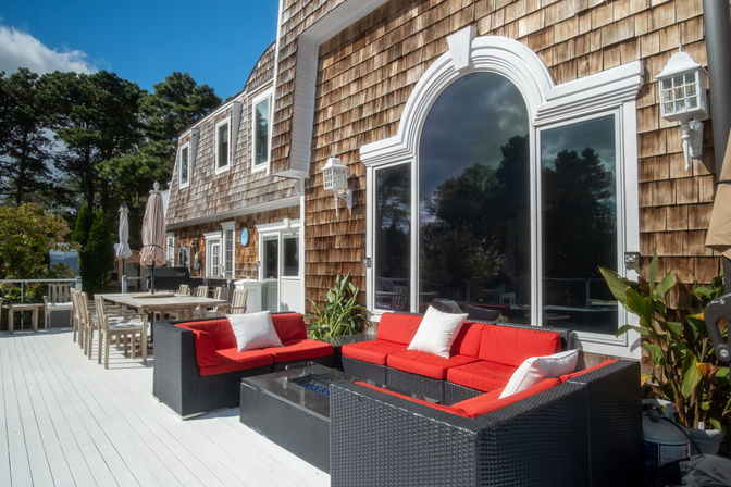 Sunny coastal shingle-style house with large arched window and white-painted deck, featuring a black wicker sectional with bright red cushions and white pillows, glass-topped firepit table, wooden dining set with umbrellas, and trees in the background.