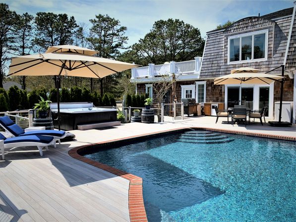 Sunny backyard pool with clear blue water and brick edge, spacious wood deck with beige cantilever umbrellas, blue cushioned lounge chairs, hot tub, outdoor dining area and grill beside a shingle-clad coastal house framed by pine trees.