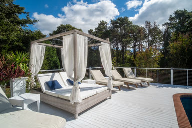Poolside white deck with a woven canopy daybed draped in sheer curtains and blue pillows, three beige sun loungers, potted tropical plants and a tree-lined backyard under a sunny sky.