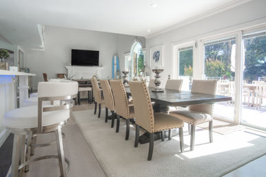 Bright modern open-concept dining room with a long dark wood table surrounded by beige nailhead chairs, white bar stools at the counter, sliding glass doors to a sunny patio, fireplace with mounted TV and a blue surfboard by the window.