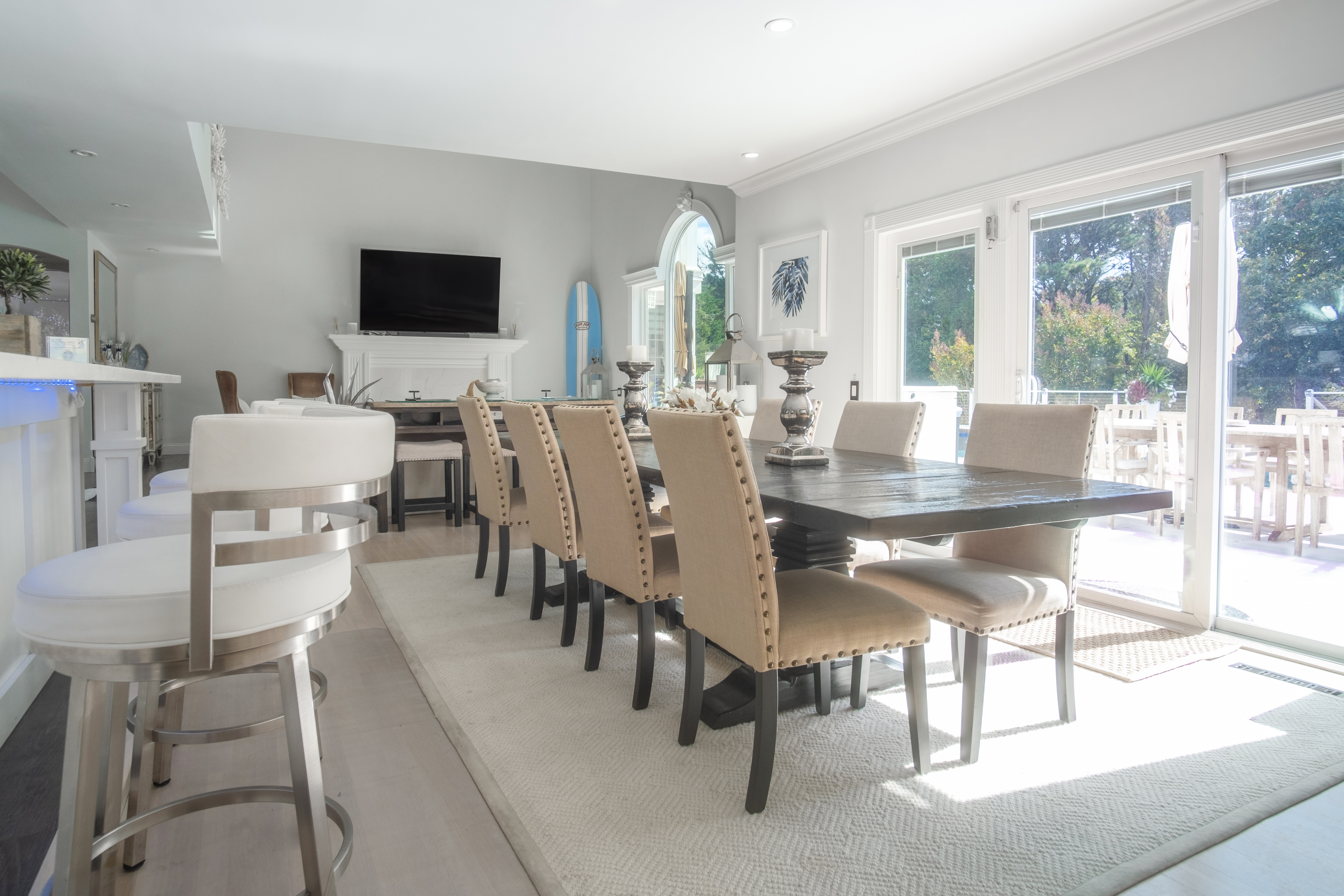 Bright modern open-concept dining room with a long dark wood table surrounded by beige nailhead chairs, white bar stools at the counter, sliding glass doors to a sunny patio, fireplace with mounted TV and a blue surfboard by the window.
