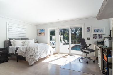 Bright sunlit coastal-style bedroom with white linens and chunky knit throw, sliding glass doors opening to a poolside deck with lounge chairs, and a compact home-office desk with an ergonomic chair.