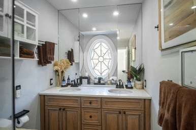 Sunlit master bathroom with double-sink wooden vanity, white marble countertop, large mirror and decorative starburst oval window, brown towels and potted plant