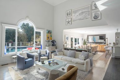 Bright coastal-inspired open living room and dining area with large arched window overlooking a backyard pool, neutral sofa, blue accent chairs, glass coffee table and kitchen island