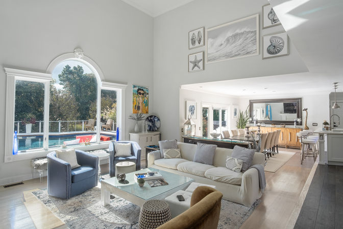 Bright coastal-inspired open living room and dining area with large arched window overlooking a backyard pool, neutral sofa, blue accent chairs, glass coffee table and kitchen island
