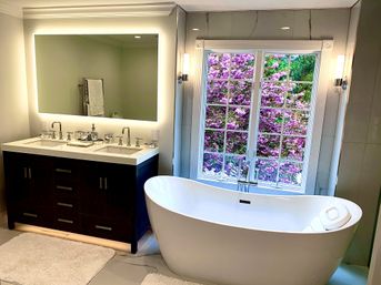 Spa-like modern bathroom with a white freestanding soaking tub by a multi-pane window showing pink flowering trees, dark wood double-sink vanity with backlit mirror and chrome fixtures on marble floor.