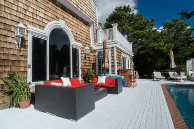 Sunny coastal shingle-style home with a white-painted pool deck, black wicker patio set with bright red cushions, potted plants, lounge chairs and umbrellas beside a blue swimming pool.