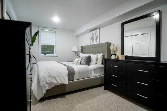 Modern minimalist bedroom interior with gray tufted upholstered bed, crisp white bedding and gray throw, black six-drawer dresser with large mirror, bedside lamp and orchid accent in a neutral carpeted room.