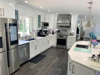 Bright modern white kitchen with stainless steel refrigerator and range, quartz island with spring-coil faucet, gray subway-tile backsplash, white shaker cabinets, pendant lights and dark wood plank floors with a vase of hydrangeas for color.