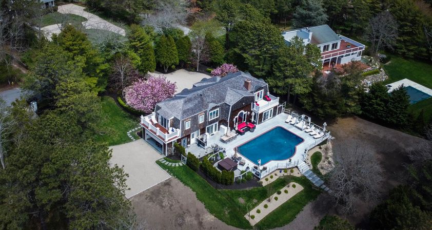 Aerial drone view of a luxury two-story home on a wooded lot with a large blue swimming pool, wraparound decks with red lounge seating, pink flowering trees and manicured gardens.