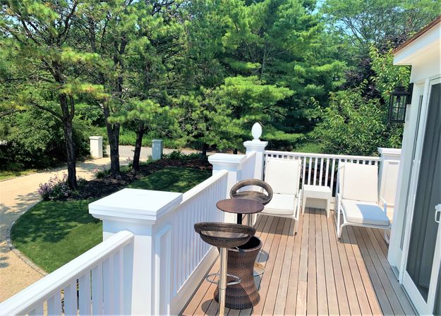 Sunny residential wooden deck with white railing, two white lounge chairs, wicker bar stools and small side table overlooking landscaped lawn, pine trees and a curved gravel driveway